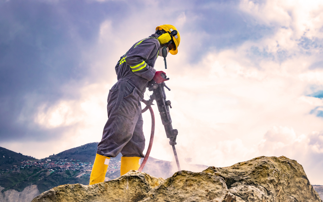 A mining worker with helmet and protective suit using a drilling machine on top of a large rock.