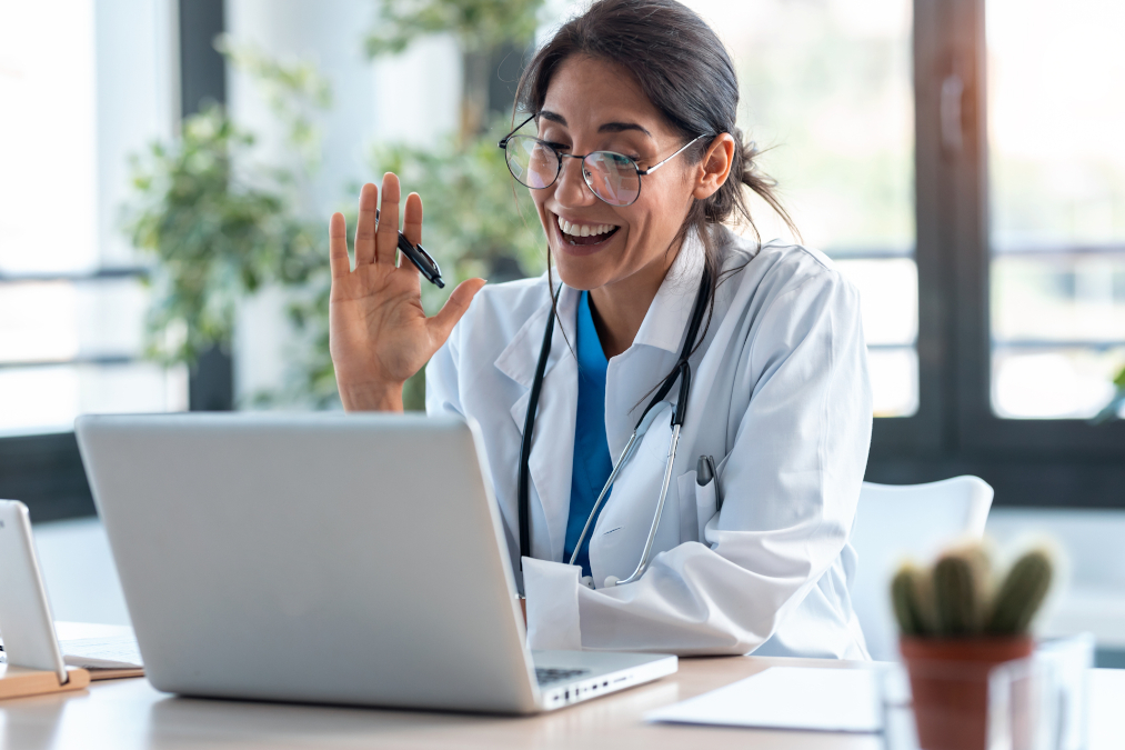 A picture of a female doctor waving and talking with colleagues through a video call with a laptop.